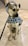A dog wearing a patterned raincoat with a hood over its head is sitting on a tiled floor. The raincoat is decorated with bone and paw prints in various colors. The dog appears to be on a leash.