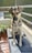 A dog with light brown and black fur is sitting on a dark cushion. Its ears are perked up, and it gazes directly at the camera. The setting is an outdoor balcony with surrounding wire mesh railing. There are potted plants nearby and wooden planks on the floor.