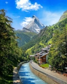 A picturesque alpine scene featuring a clear blue stream flowing alongside a road lined with charming chalet-style buildings. Tall green trees frame the image on either side, with a majestic, snow-capped mountain towering in the background under a bright blue sky with fluffy white clouds.