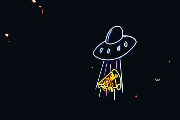 Neon lights create the image of a UFO beaming up a slice of pizza against a dark background.