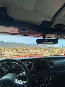 The view from inside a vehicle shows desert scenery with dry shrubs and a mountain range in the background. A few people are walking along a wooden fence, and a yellow traffic sign is visible beside them.