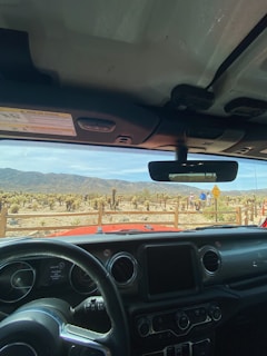 The view from inside a vehicle shows desert scenery with dry shrubs and a mountain range in the background. A few people are walking along a wooden fence, and a yellow traffic sign is visible beside them.
