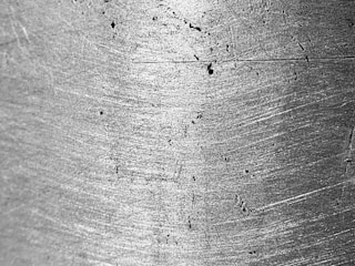 A close-up view of a metallic surface with visible scratches and wear marks. The texture is rough, with light reflecting off its uneven surface, highlighting the grain and imperfections.