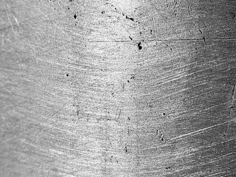 A close-up view of a metallic surface with visible scratches and wear marks. The texture is rough, with light reflecting off its uneven surface, highlighting the grain and imperfections.
