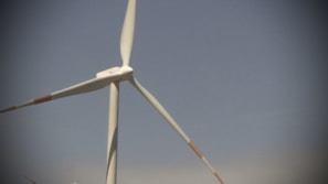 A large wind turbine with three blades set against a clear blue sky, with parts of other turbines visible in the background.