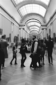 A black and white photograph capturing a spacious art gallery with a high, arched ceiling and several people walking and observing the art. The gallery walls feature numerous framed paintings, and the floor is made of large, patterned tiles.