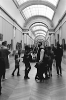 A black and white photograph capturing a spacious art gallery with a high, arched ceiling and several people walking and observing the art. The gallery walls feature numerous framed paintings, and the floor is made of large, patterned tiles.