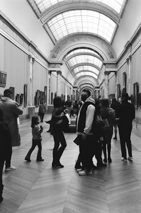 A black and white photograph capturing a spacious art gallery with a high, arched ceiling and several people walking and observing the art. The gallery walls feature numerous framed paintings, and the floor is made of large, patterned tiles.