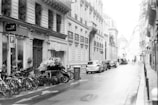 A black and white photograph of a European city street, adorned with classical architecture. Several bicycles are parked on the left side near a flower shop with a variety of plants and bouquets on display. Cars are lined up along the cobblestone road, and a few pedestrians can be seen walking in the distance.