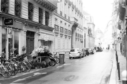 A black and white photograph of a European city street, adorned with classical architecture. Several bicycles are parked on the left side near a flower shop with a variety of plants and bouquets on display. Cars are lined up along the cobblestone road, and a few pedestrians can be seen walking in the distance.