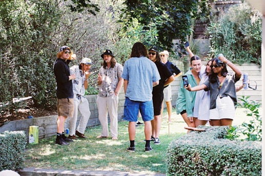 A group of people gathers outdoors in a lush garden setting. Several individuals are casually dressed, with some holding drinks. Two people in the foreground appear to be taking a selfie together. The atmosphere is relaxed and social, with greenery and wooden elements visible in the background.