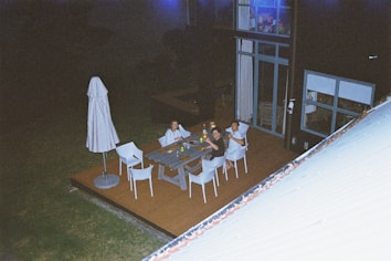Three people are seated around a rectangular outdoor table on a patio at night. The patio is attached to a modern building with large windows. There are several chairs around the table and an umbrella on the patio. The atmosphere is relaxed as the group seems to be enjoying drinks and conversation.