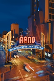 The image depicts a vibrant night scene in an urban environment, showcasing a large illuminated sign that reads 'Reno' and 'The Biggest Little City in the World' in bright neon lights. The sign spans across a street with light trails from moving vehicles creating dynamic streaks on the road. Tall buildings with lit windows are visible in the background.