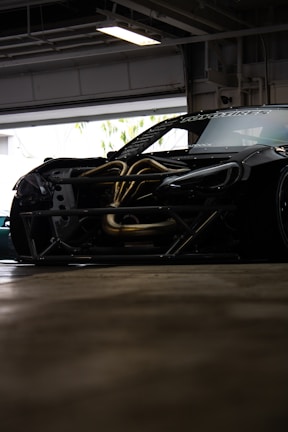 A sleek, modified sports car with exposed engine components is parked inside a garage. The vehicle has a dark exterior with visible aerodynamic elements, such as a significant body kit. Parts of the hood are removed, revealing intricately designed metal pipes and components.