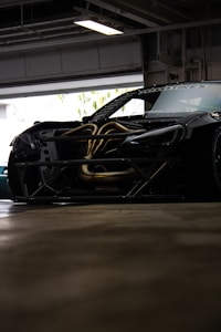 A sleek, modified sports car with exposed engine components is parked inside a garage. The vehicle has a dark exterior with visible aerodynamic elements, such as a significant body kit. Parts of the hood are removed, revealing intricately designed metal pipes and components.