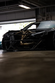 A sleek, modified sports car with exposed engine components is parked inside a garage. The vehicle has a dark exterior with visible aerodynamic elements, such as a significant body kit. Parts of the hood are removed, revealing intricately designed metal pipes and components.