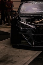 A close-up view of a sleek, modified black car with visible branding and decals on the windshield. Part of the car's engine is exposed, showcasing intricate mechanical details. In the background, several people are standing, suggesting an event or gathering.