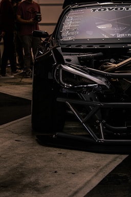 A close-up view of a sleek, modified black car with visible branding and decals on the windshield. Part of the car's engine is exposed, showcasing intricate mechanical details. In the background, several people are standing, suggesting an event or gathering.