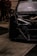 A close-up view of a sleek, modified black car with visible branding and decals on the windshield. Part of the car's engine is exposed, showcasing intricate mechanical details. In the background, several people are standing, suggesting an event or gathering.