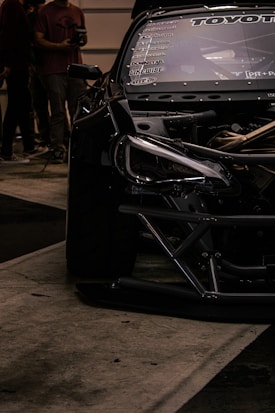 A close-up view of a sleek, modified black car with visible branding and decals on the windshield. Part of the car's engine is exposed, showcasing intricate mechanical details. In the background, several people are standing, suggesting an event or gathering.