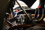 A close-up view of a car engine with visible metallic parts and intricate piping. The background is slightly blurred, showing people and another vehicle.