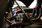 A close-up view of a car engine with visible metallic parts and intricate piping. The background is slightly blurred, showing people and another vehicle.