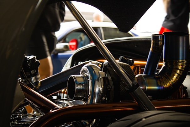 A close-up view of a car engine with visible metallic parts and intricate piping. The background is slightly blurred, showing people and another vehicle.
