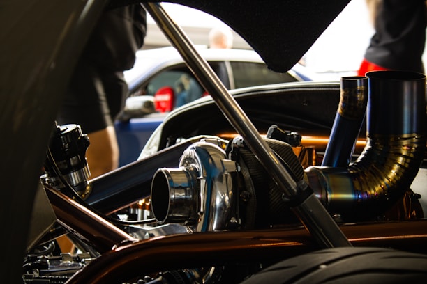 A close-up view of a car engine with visible metallic parts and intricate piping. The background is slightly blurred, showing people and another vehicle.