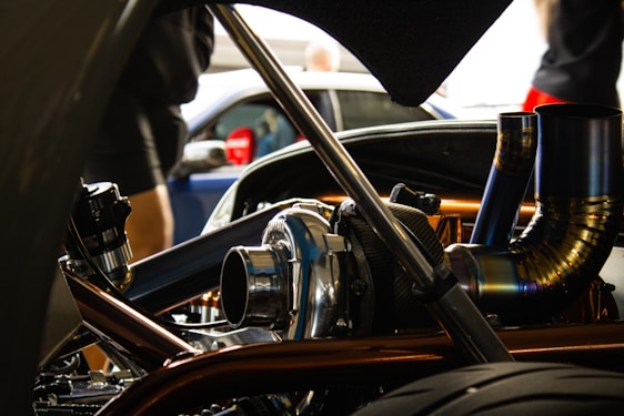 A close-up view of a car engine with visible metallic parts and intricate piping. The background is slightly blurred, showing people and another vehicle.