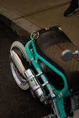 A custom motorcycle with a prominent turquoise frame and a luxurious brown leather seat, showcasing intricate designs with a patterned texture. The chrome exhaust and various mechanical components are polished and reflective. A visible footpath and part of a person's shoe are in the background.