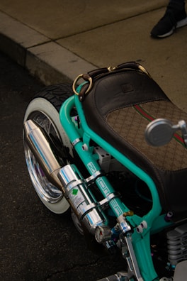 A custom motorcycle with a prominent turquoise frame and a luxurious brown leather seat, showcasing intricate designs with a patterned texture. The chrome exhaust and various mechanical components are polished and reflective. A visible footpath and part of a person's shoe are in the background.
