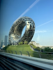 A large, futuristic silver structure with intricate black inscriptions stands prominently on a grassy hill. The structure resembles a torus and is positioned against a clear blue sky, with modern buildings visible in the background.