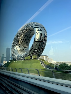 A large, futuristic silver structure with intricate black inscriptions stands prominently on a grassy hill. The structure resembles a torus and is positioned against a clear blue sky, with modern buildings visible in the background.
