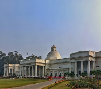 A large white neoclassical building with a prominent dome and columns, surrounded by a manicured garden and pathways. The building features an Indian national flag and inscriptions in Hindi on the facade. The sky is clear with a soft, diffused light.