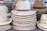 A stack of straw hats with varying woven patterns and colors, some featuring leather bands around the crown. The hats are neatly piled and organized in rows, creating a textured and symmetrical display.