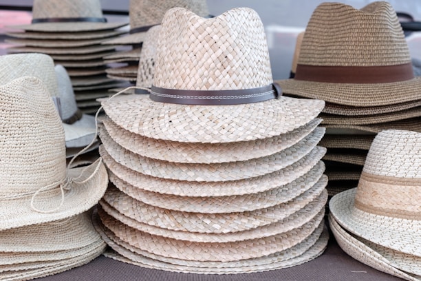 A stack of straw hats with varying woven patterns and colors, some featuring leather bands around the crown. The hats are neatly piled and organized in rows, creating a textured and symmetrical display.