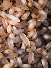 A close-up view of uncooked rice grains, displaying a combination of medium to long grain types with slight brown streaks and imperfections. The grains have a translucent quality and are closely packed together.