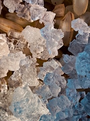 A close-up view of translucent, crystal-like structures alongside pale brown, elongated grains. The crystals have intricate facets that catch and reflect light, creating a sparkling effect. The grains underneath have a subtle texture, contrasting with the clear, icy appearance of the crystals.