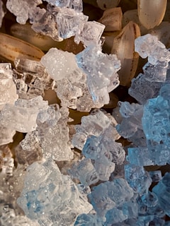 A close-up view of translucent, crystal-like structures alongside pale brown, elongated grains. The crystals have intricate facets that catch and reflect light, creating a sparkling effect. The grains underneath have a subtle texture, contrasting with the clear, icy appearance of the crystals.
