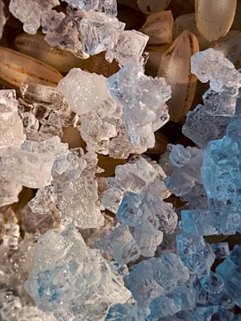 A close-up view of translucent, crystal-like structures alongside pale brown, elongated grains. The crystals have intricate facets that catch and reflect light, creating a sparkling effect. The grains underneath have a subtle texture, contrasting with the clear, icy appearance of the crystals.