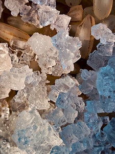 A close-up view of translucent, crystal-like structures alongside pale brown, elongated grains. The crystals have intricate facets that catch and reflect light, creating a sparkling effect. The grains underneath have a subtle texture, contrasting with the clear, icy appearance of the crystals.