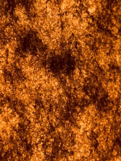 The image features a textured surface with a predominantly orange and black color palette, providing a rough and granular appearance. The surface has an organic, irregular pattern that mimics natural formations or material fibers.