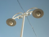 A tall, white street lamp pole with two lights extends upwards against a clear blue sky. The design is modern, featuring curved elements near the light fixtures. An electrical wire runs diagonally across the image.