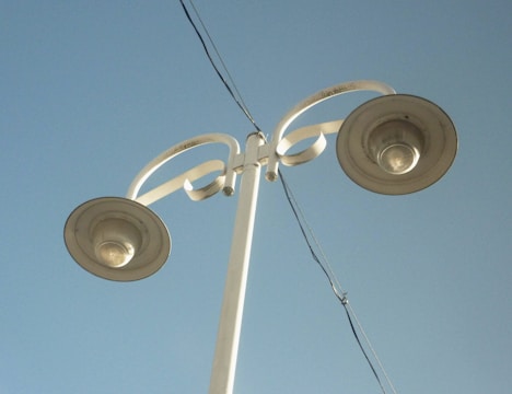 A tall, white street lamp pole with two lights extends upwards against a clear blue sky. The design is modern, featuring curved elements near the light fixtures. An electrical wire runs diagonally across the image.
