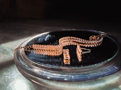 A gold-colored, intricately designed jewelry piece is resting on a reflective surface. The jewelry features multiple rectangular links and is likely a bracelet or necklace. It is displayed in soft lighting, emphasizing its shine and texture.