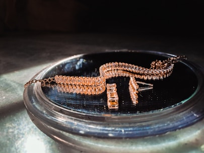 A gold-colored, intricately designed jewelry piece is resting on a reflective surface. The jewelry features multiple rectangular links and is likely a bracelet or necklace. It is displayed in soft lighting, emphasizing its shine and texture.
