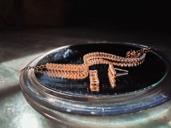 A gold-colored, intricately designed jewelry piece is resting on a reflective surface. The jewelry features multiple rectangular links and is likely a bracelet or necklace. It is displayed in soft lighting, emphasizing its shine and texture.