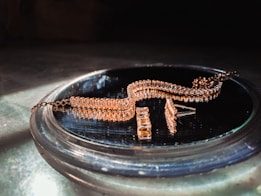 A gold-colored, intricately designed jewelry piece is resting on a reflective surface. The jewelry features multiple rectangular links and is likely a bracelet or necklace. It is displayed in soft lighting, emphasizing its shine and texture.