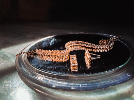 A gold-colored, intricately designed jewelry piece is resting on a reflective surface. The jewelry features multiple rectangular links and is likely a bracelet or necklace. It is displayed in soft lighting, emphasizing its shine and texture.