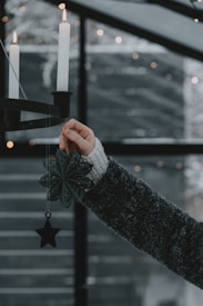 A hand wearing a dark knitted sweater holds a decorative metal snowflake and star hanging from a candle holder. Two white candles are lit and the background features a dimly lit, cozy space with small string lights in a blurred pattern.
